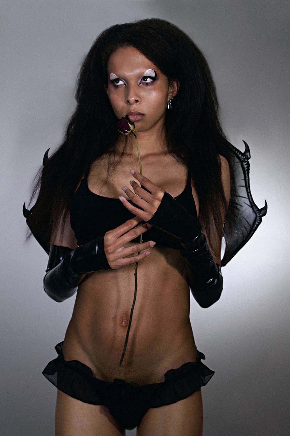 Alessa facing the camera with graphic eyeliner wearing black lingere, latex gloves and bat wings on a gray background